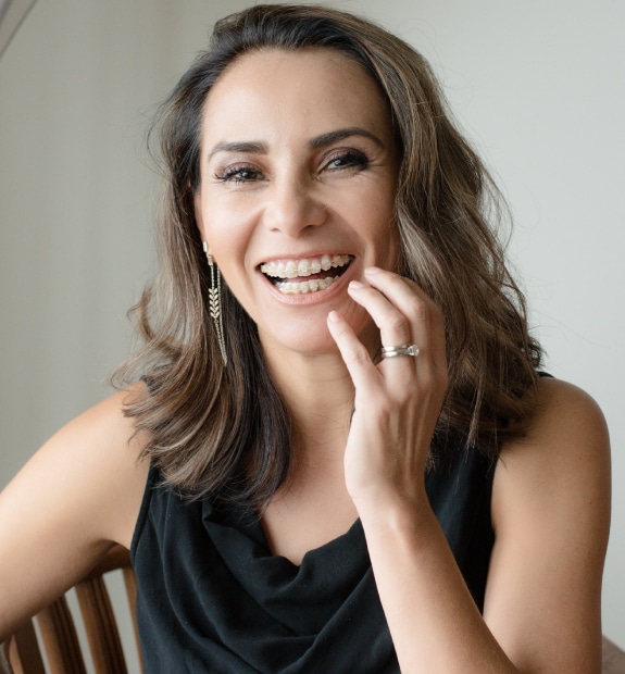 Woman smiling while touching her face, wearing black sleeveless top against neutral background.
