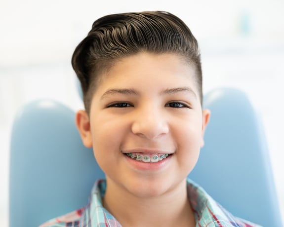 Young boy smiling at camera in orthodontist's office with blue chair visible behind him.