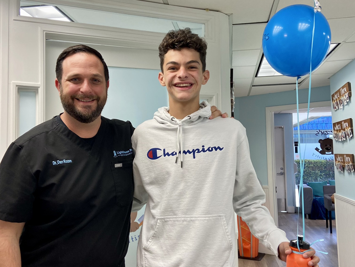 Two men posing together in an orthodontic office with a blue balloon visible in the background.