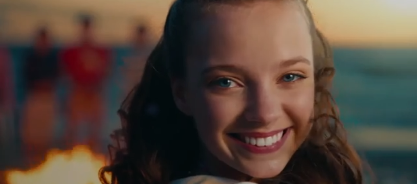 Young girl smiling at camera with beach and sunset in blurred background.