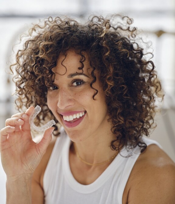 Woman with curly brown hair smiling while holding an Invisalign aligner tray.