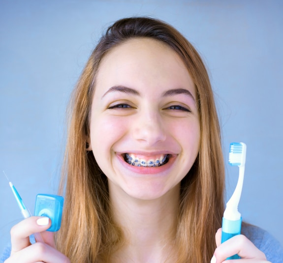 Smiling teenager with braces holding toothbrushes against blue background for orthodontics website.