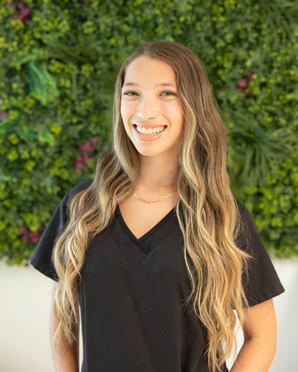 Bella smiles at the camera wearing a black v-neck shirt in front of a green plant wall.